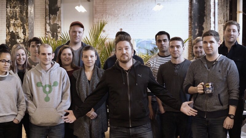 Group of colleagues gathered in an exposed-brick office, a man in a black hoodie at the front with arms held out