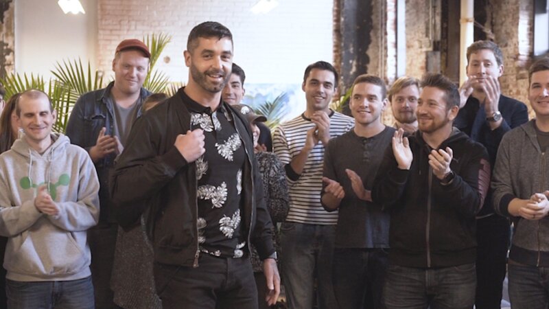 Colleagues applauding in an exposed-brick office as a smiling man in a dark jacket looks on